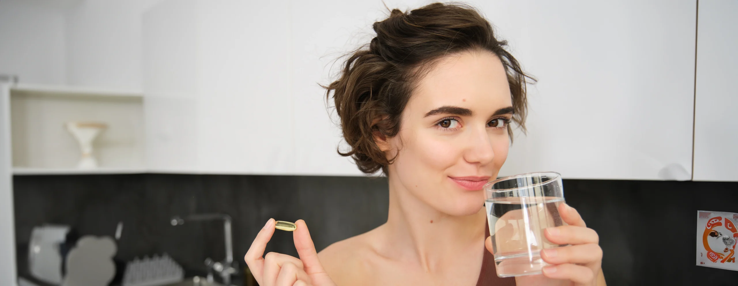 kobieta trzyma kapsułkę z omega 3 na długowieczność w palcach, w drugiej dłoni szklankę z wodą, aby popić kapsułkę